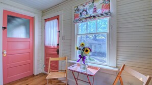 A room with a pink door, a window with a decorative valance, a wooden chair, a small table with a vase of flowers, and a wooden bench.