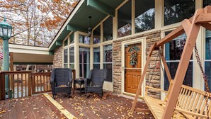 Une terrasse en bois avec des chaises et une balançoire, un mur en pierre et une porte ornée d’une guirlande.