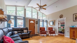 Un salon spacieux avec un canapé en cuir, deux fauteuils à motifs, une table basse en bois et un ventilateur de plafond. De grandes fenêtres laissent entrer la lumière naturelle et offrent une vue sur l’extérieur.