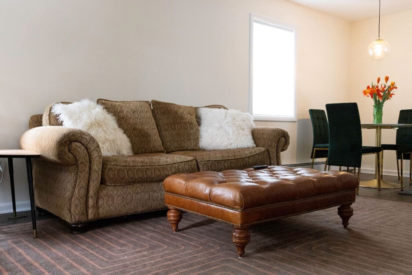 Een woonkamer met een bruine bank, witte zachte kussens, een bruin leren poef, een klein bijzettafeltje, en een eethoek met groene stoelen en een bloevas.
