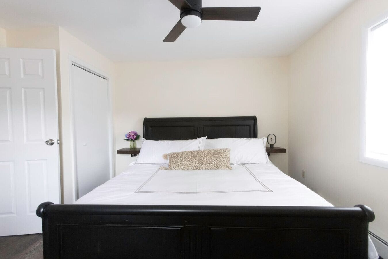 A bedroom with a bed, a ceiling fan, a nightstand with a vase of flowers, and a window.