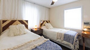 Habitación de hotel con dos camas, ventilador de techo, mesita de noche con lámpara y ventana con vistas.