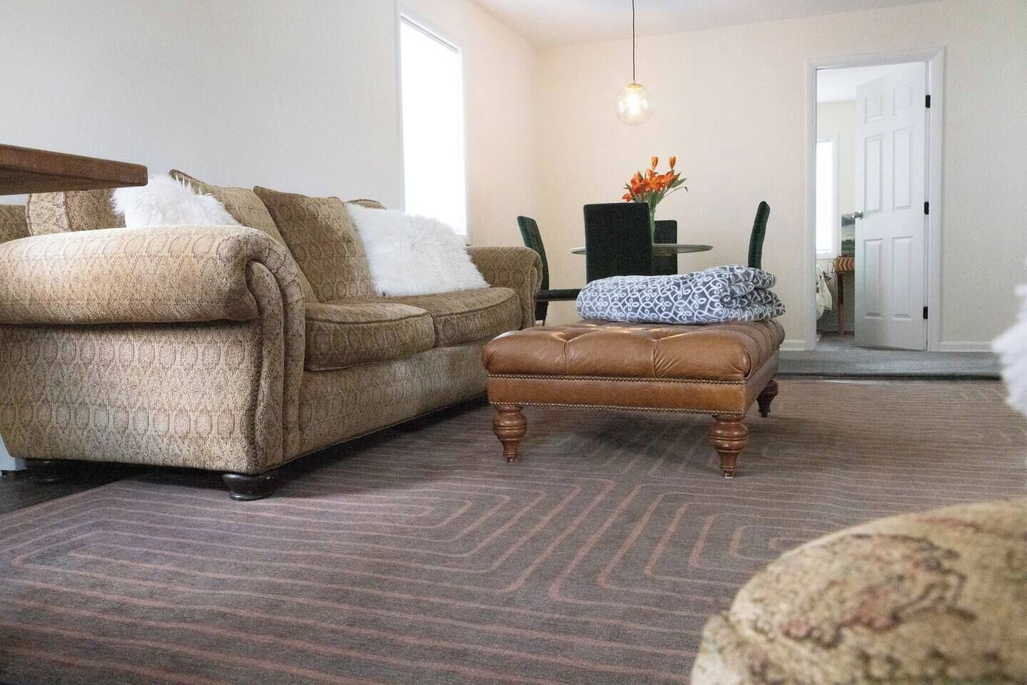 Ein Wohnzimmer mit braunem Ledersofa, passendem Hocker, einem gemusterten Teppich und einem Essbereich mit Tisch und Stühlen.