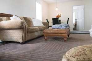 A living room with a brown leather sofa, a matching ottoman, a patterned rug, and a dining area with a table and chairs.