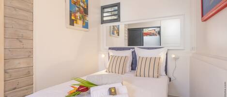 A neatly made bed with white linens, a small table with a plant, and a painting on the wall.