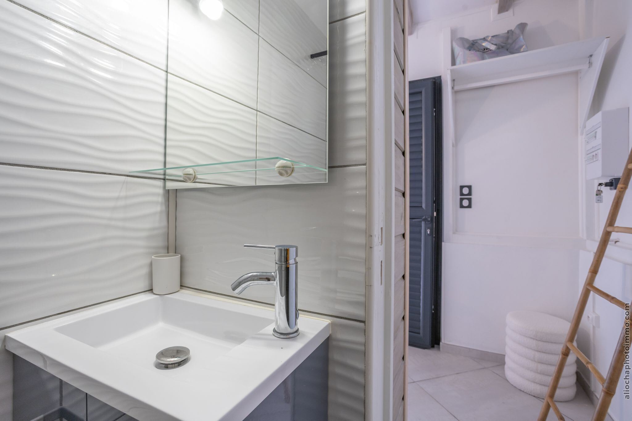 A modern bathroom with a white sink, chrome faucet, and a glass shelf above the mirror.