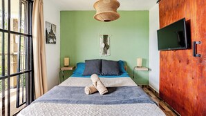 A bedroom with a bed, a television, a macrame hanging, and a view through a window.