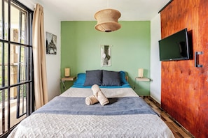 Un dormitorio con cama, televisor, un colgador de macramé y vistas al exterior a través de una ventana.