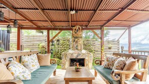 A covered outdoor seating area with a stone fireplace, wooden furniture, and a view of the outdoors.