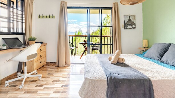A bedroom with a bed, a desk with a laptop, a chair, a balcony with chairs, and a view of the outdoors.