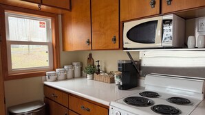 A compact kitchen with wooden cabinets, a microwave, a stove, and a window with a view of a grassy area.