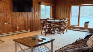 A cozy cabin interior with a wooden floor, a dining area, a TV mounted on the wall, and a ceiling fan.