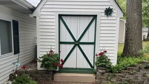 A white shed with a green door and a cross design, surrounded by plants and a stone wall.