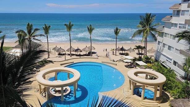 A resort pool with a circular design, surrounded by palm trees and beach umbrellas, overlooking a beach and ocean. - 2-bedroom apartment in peaceful Mazatlán with an amazing front ocean view (Mazatlán)