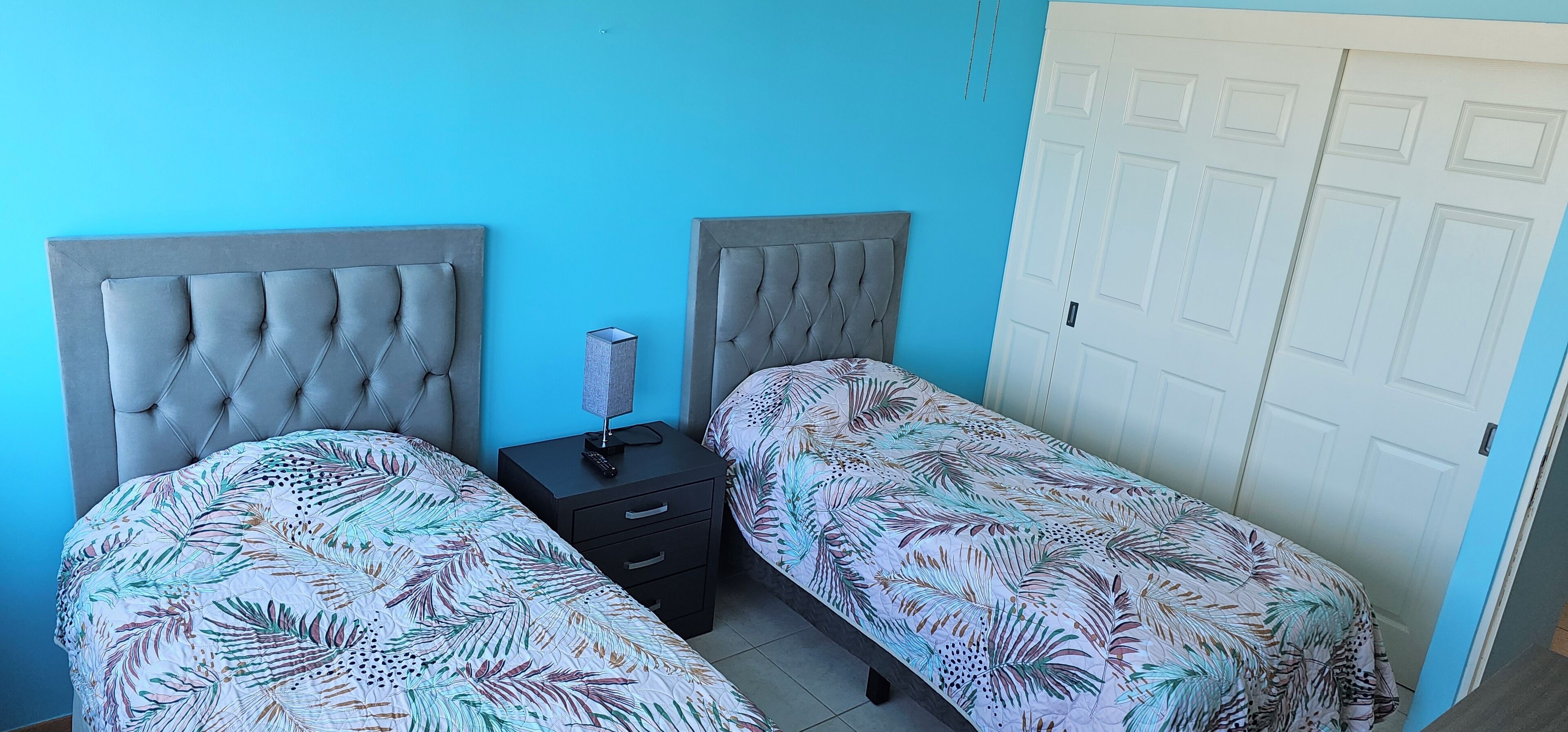 Une chambre avec deux lits dotés de têtes de lit capitonnées, une table de chevet avec une lampe et une porte blanche.