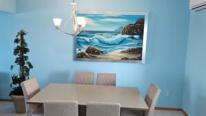 A dining area with a table and chairs, a chandelier, a potted plant, and a painting of a coastal scene. - 2-bedroom apartment in peaceful Mazatlán with an amazing front ocean view (Mazatlán)