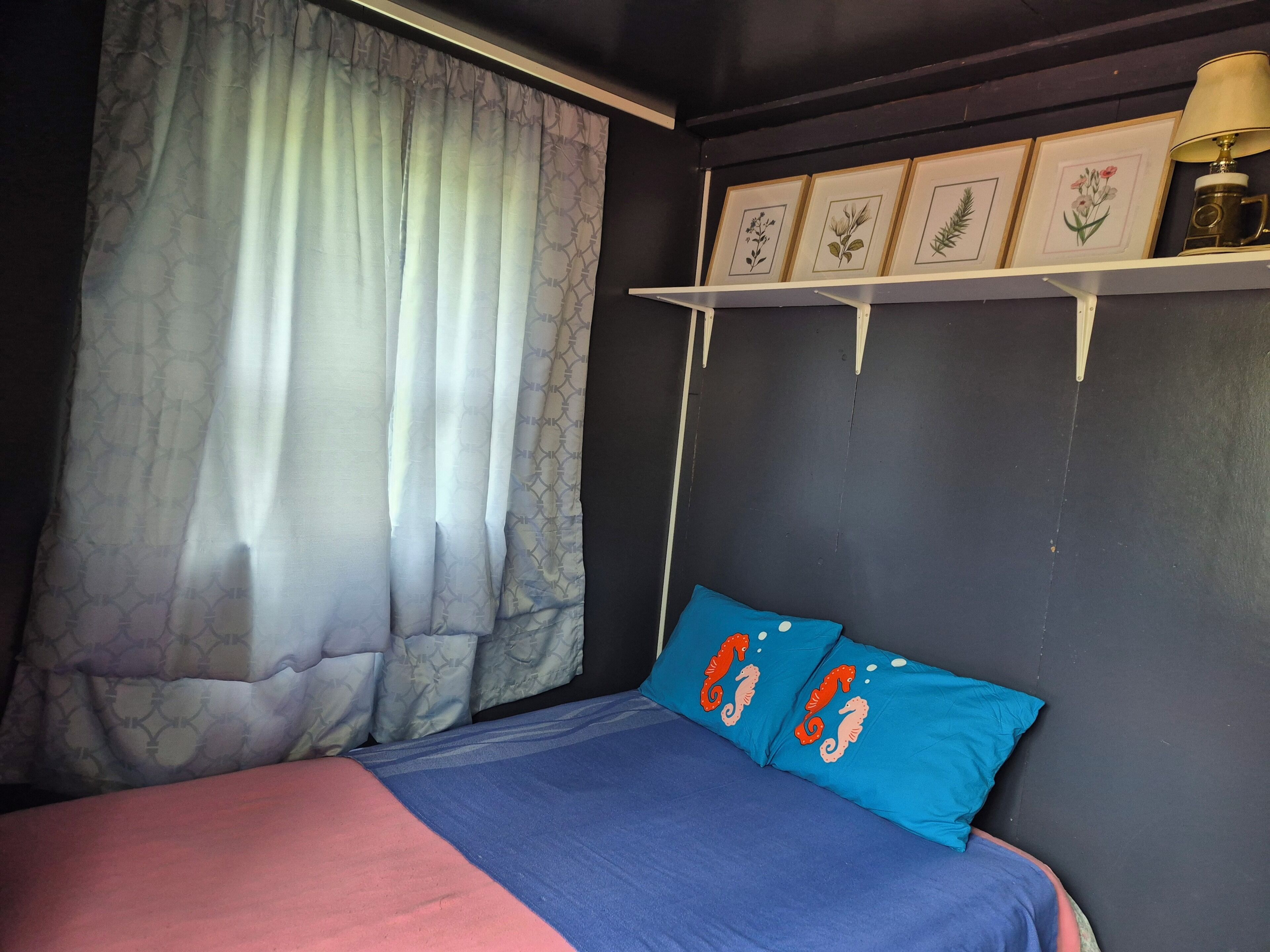 A bedroom with a bed, a window with curtains, a wall-mounted shelf with framed pictures, and a lamp.