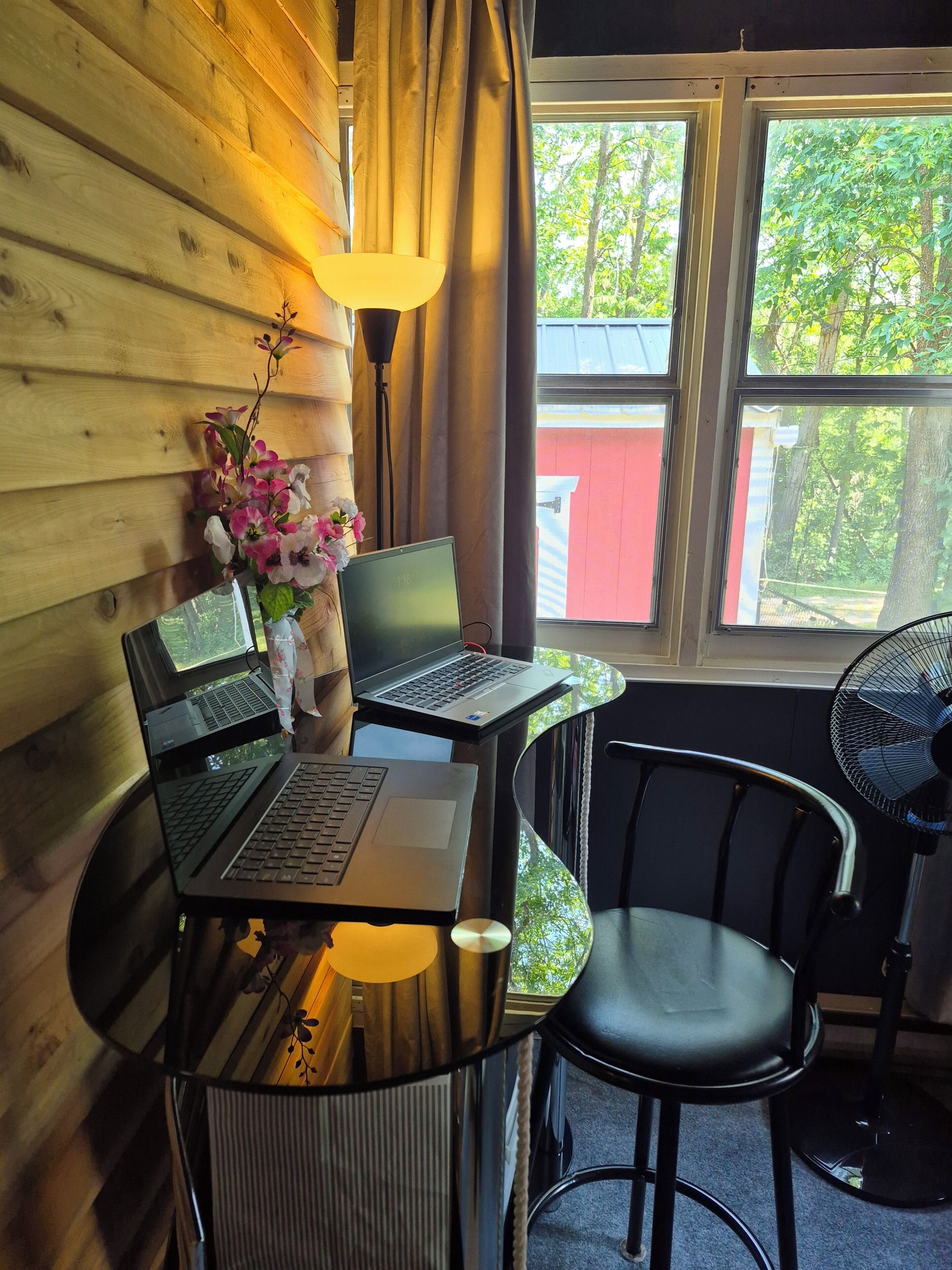 A room with two laptops on a glass table, a chair, a lamp, and a view of greenery outside.