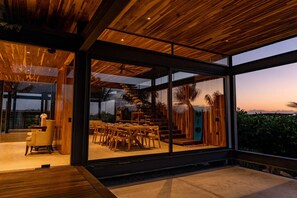 Un interior moderno con grandes ventanales, techo de madera y una zona de comedor con sillas y mesas.