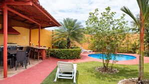 Ein überdachter Außenbereich mit Tischen und Stühlen, ein Schwimmbecken und ein kleiner Garten mit einer Palme und Sträuchern.