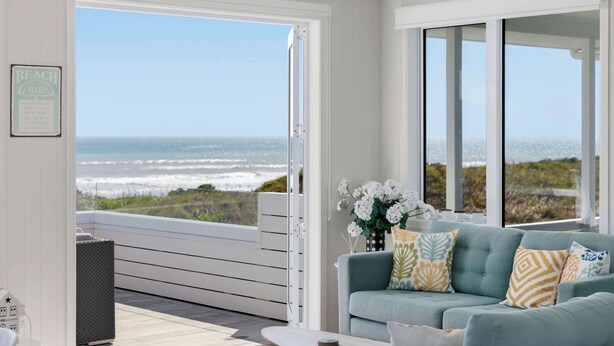 Una stanza con un'ampia porta finestra scorrevole che dà su un balcone con vista sull'oceano. Si vede un divano azzurro con cuscini decorativi e un vaso di fiori bianchi.