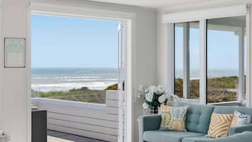 A room with a large sliding glass door leading to a balcony with a view of the ocean. There is a light blue sofa with decorative pillows and a vase of white flowers.