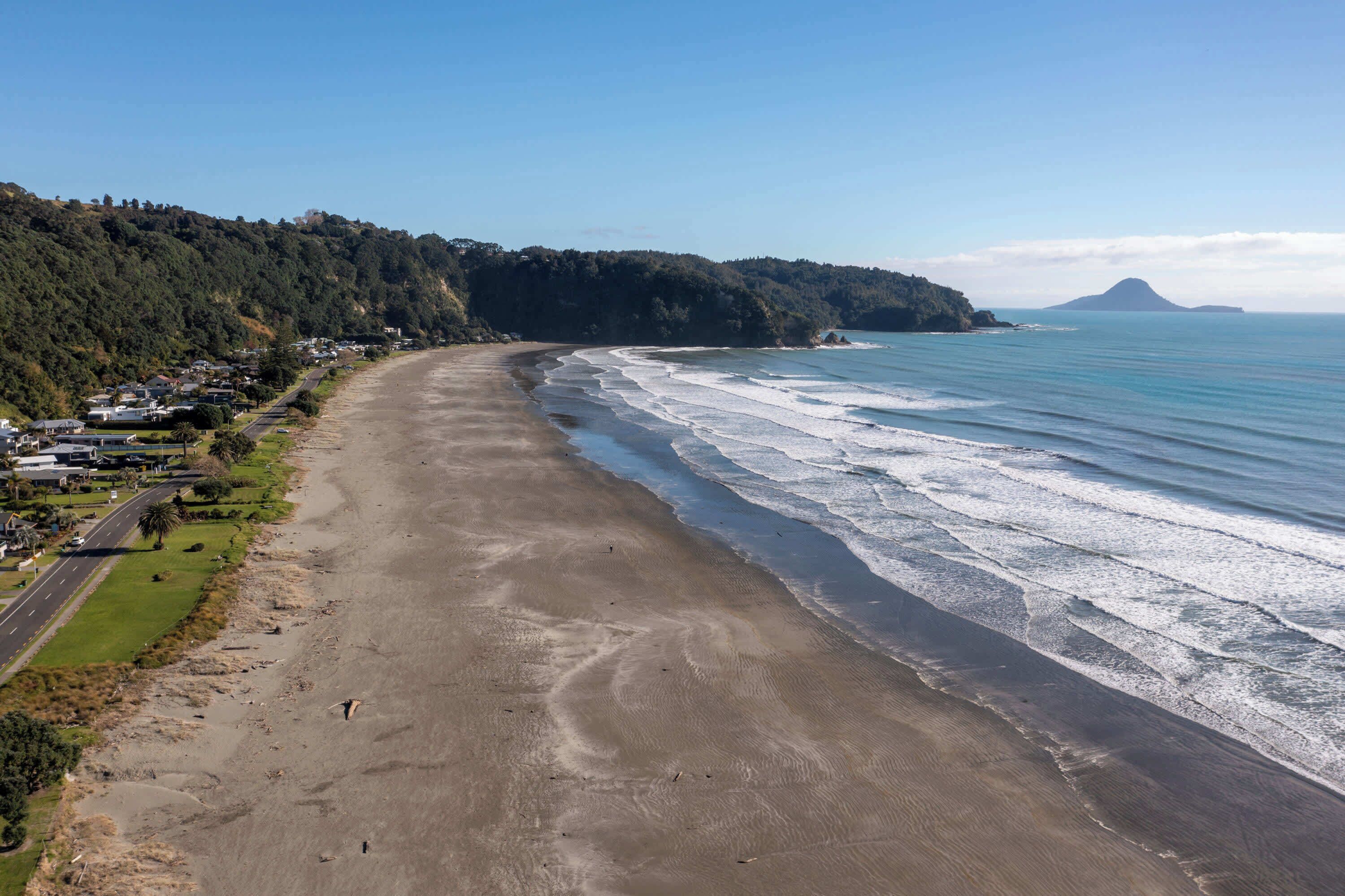 Una playa costera con una carretera que recorre la orilla, una colina boscosa al fondo y un cielo despejado.