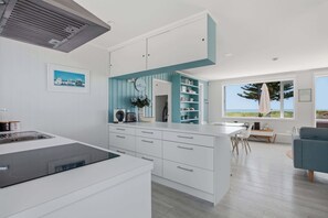 A modern kitchen with white cabinetry, a built-in island, and a view of the ocean through large windows. - Ohope Beach Bliss Stunning Sea Views & Family fun (Ōhope)
