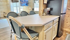 Interior de una cabaña de madera con una cocina que incluye una isla grande, sillas, un refrigerador y una cafetera.