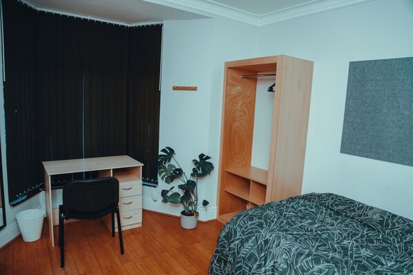 A bedroom with a wooden desk, a black chair, a bed with a leaf-patterned bedspread, a potted plant, and a closet. - The Phoenix (Swansea)