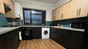 A compact kitchen with wooden cabinets, a built-in washing machine, and a window with a view of buildings. - The Phoenix  (Swansea)