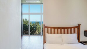 Ein Schlafzimmer mit einem hölzernen Kopfteil, einem großen Fenster mit Blick auf das Meer und Bäume sowie einer weißen Bettdecke.