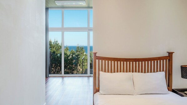 Une chambre à coucher avec une tête de lit en bois, une grande fenêtre donnant sur l’océan et les arbres, et un couvre-lit blanc.