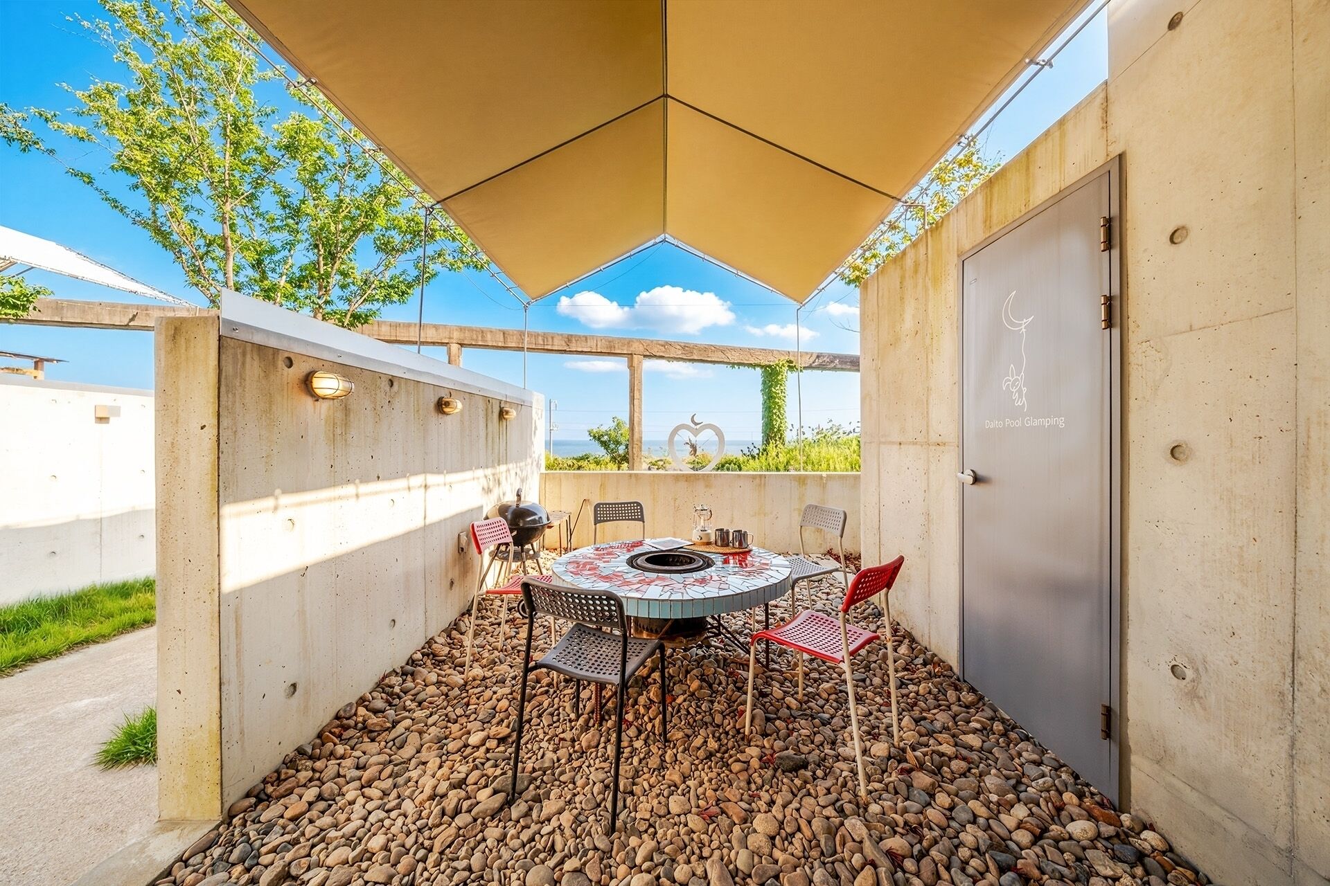 A covered outdoor seating area with a table and chairs, a grill, and a view of the sea.