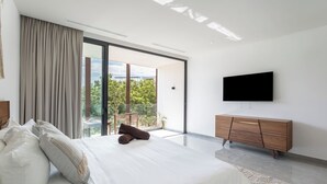 Ein modernes Schlafzimmer mit einem großen Bett, einer Holzkommode, einem Flachbildfernseher und einem Balkon mit Blick auf Grünflächen.