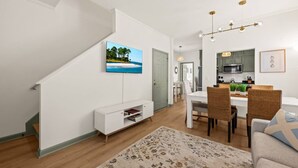 A modern living room with a flat-screen TV, a dining area with wicker chairs, a kitchen with green cabinets, and a staircase. - Charleston 3BR Home · 3 Baths · Porches · Dogs OK (North Charleston)