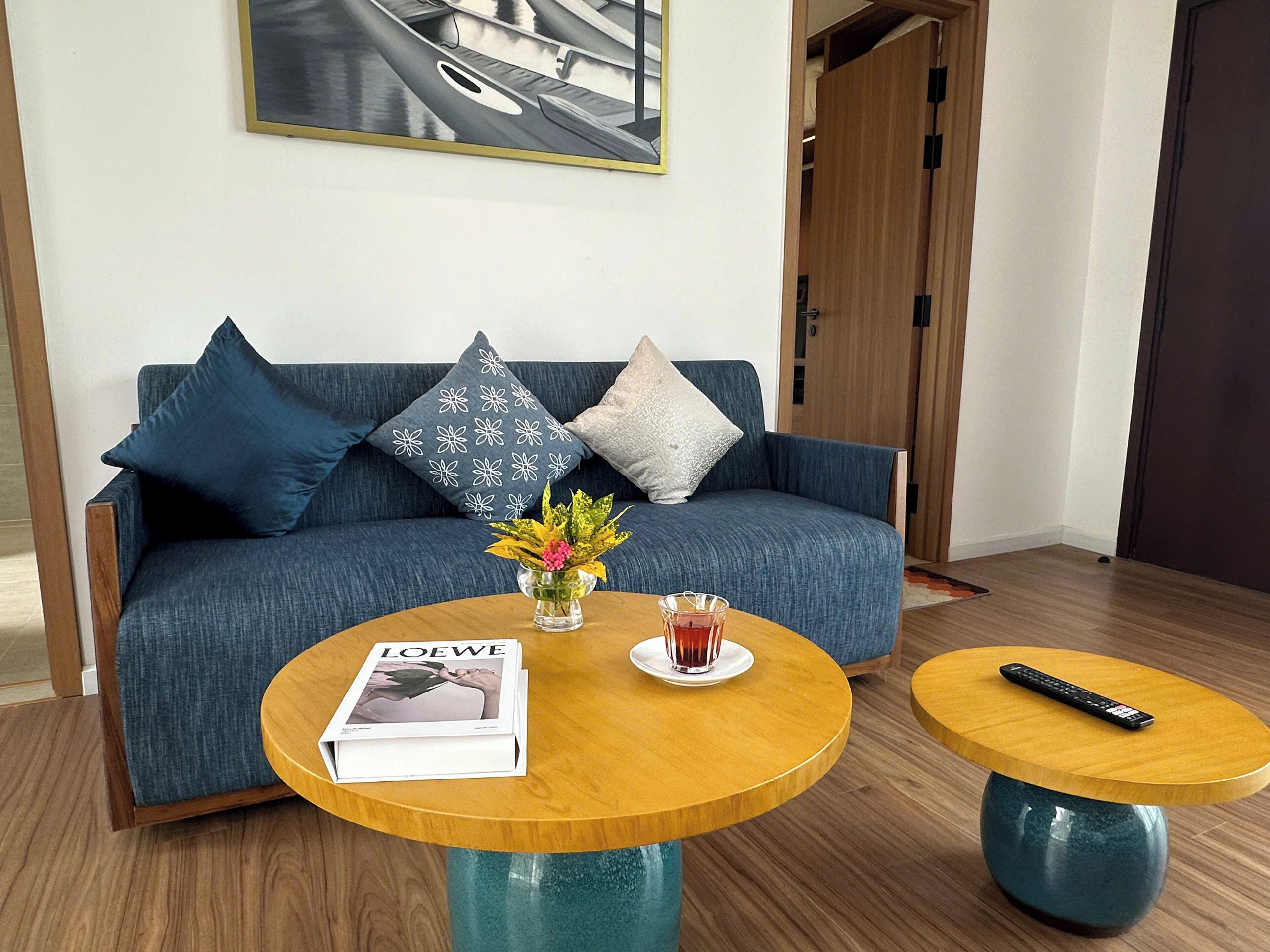 A modern living room with a blue sofa, two wooden side tables, a magazine, a vase with flowers, and a remote control.