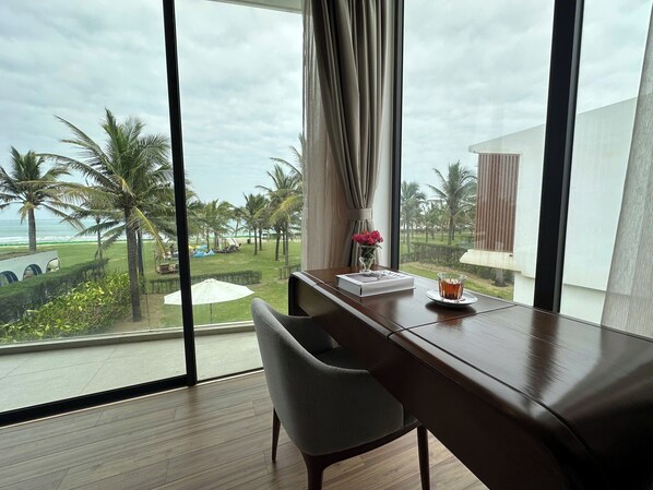 A room with a large window overlooking a landscaped area with palm trees and a view of the ocean. There is a desk with a chair, a vase with flowers, and a glass of water on the windowsill. - Hoi An Ocean Villas (Hoi An)