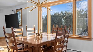 Ein Essbereich mit Holztisch und Stühlen, einer modernen Chandelier und großen Fenstern mit Blick auf Bäume und Himmel.