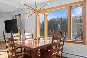 A dining area with a wooden table and chairs, a modern chandelier, and large windows offering a view of trees and sky. - Centrally Located, Renovated 4BR Stowe Getaway! (Stowe)