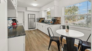 A modern kitchen with white cabinets, a stainless steel refrigerator, a granite countertop, and a dining area with a white table and black chairs. - Coastal Retreat w/ Heated Pool & Games (Largo)