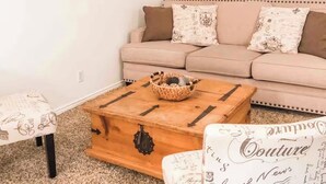 A cozy living room with a beige sofa, a wooden coffee table with a basket, a small table with a cushion, and a chair with a printed cushion.
