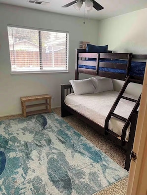 Una habitación con literas, un banco de madera pequeño, un tapete estampado y una ventana con persianas.