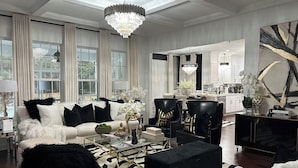 Ein geräumiges Wohnzimmer mit einem weißen Sofa, schwarzen Ohrensesseln, einem Glastisch und einem schwarzen Hocker. Eine Kristallleuchter hängt von der Decke, und es gibt ein großes Fenster mit Vorhängen.