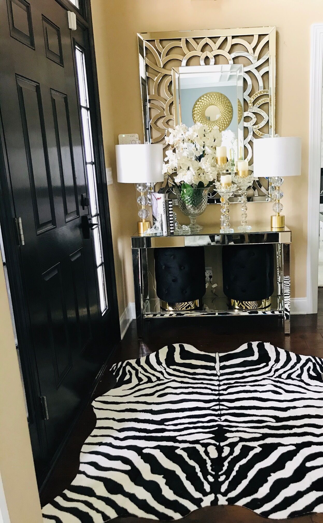 Une porte noire, un miroir encadré d’or, une console noire et dorée agrémentée de lampes blanches, et un tapis à rayures zébrées.