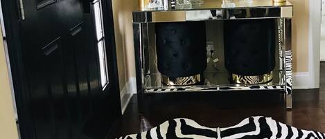 Une porte noire, un miroir encadré d’or, une console noire et dorée agrémentée de lampes blanches, et un tapis à rayures zébrées.