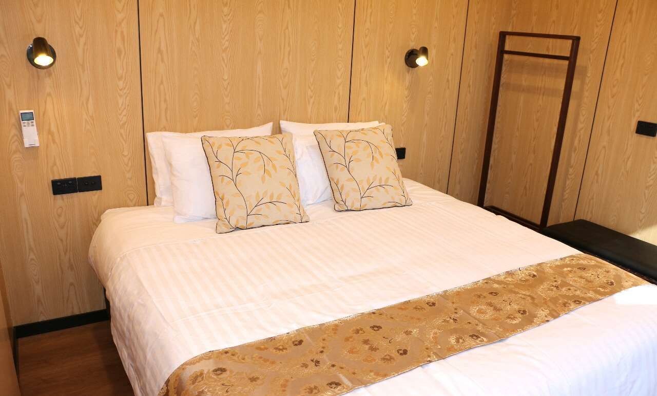 A hotel room with a wooden headboard, a bed with white bedding and two decorative pillows, a bedside table, and a wall-mounted control panel.
