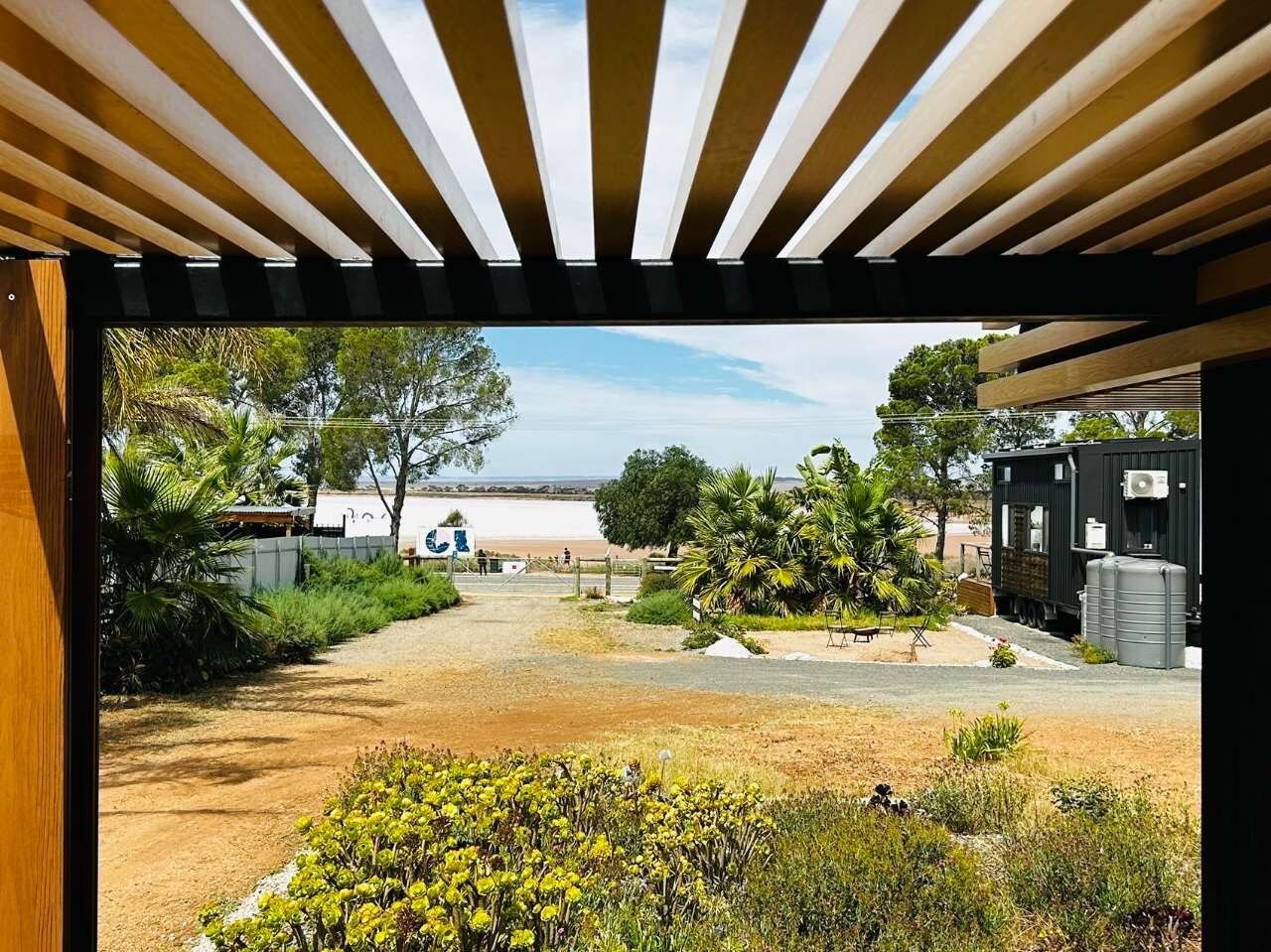 Blick durch ein schmales Holsterfenster auf einen Strand, mit einem Kiesweg, der zu einem schwarzen Gebäude mit Wassertank führt.