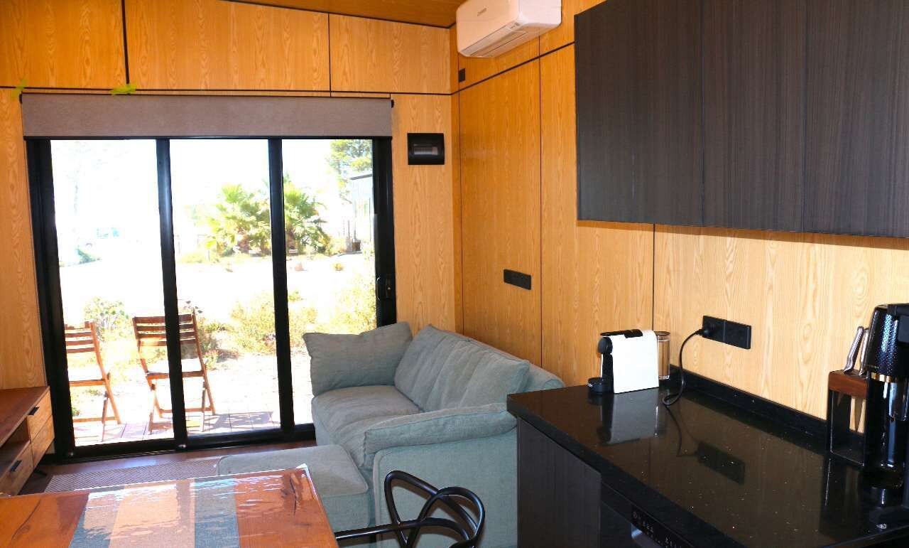 A compact living space with a sofa, a coffee machine, and a sliding glass door leading to an outdoor area with chairs.