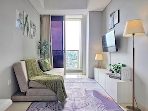 A modern living room with a sofa, a TV, a lamp, and a view of the outdoors. - Arumi By SanLiving - 2BR - Direct Mall (Jakarta)